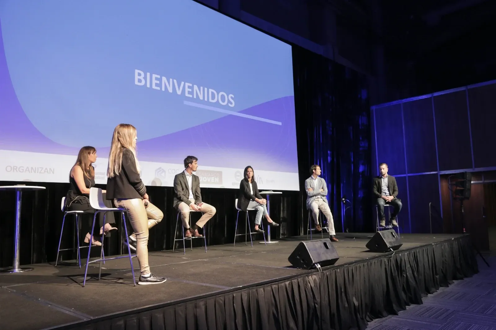 Panel sobre el escenario, con la pantalla de bienvenida detrás y los monitores de piso al frente