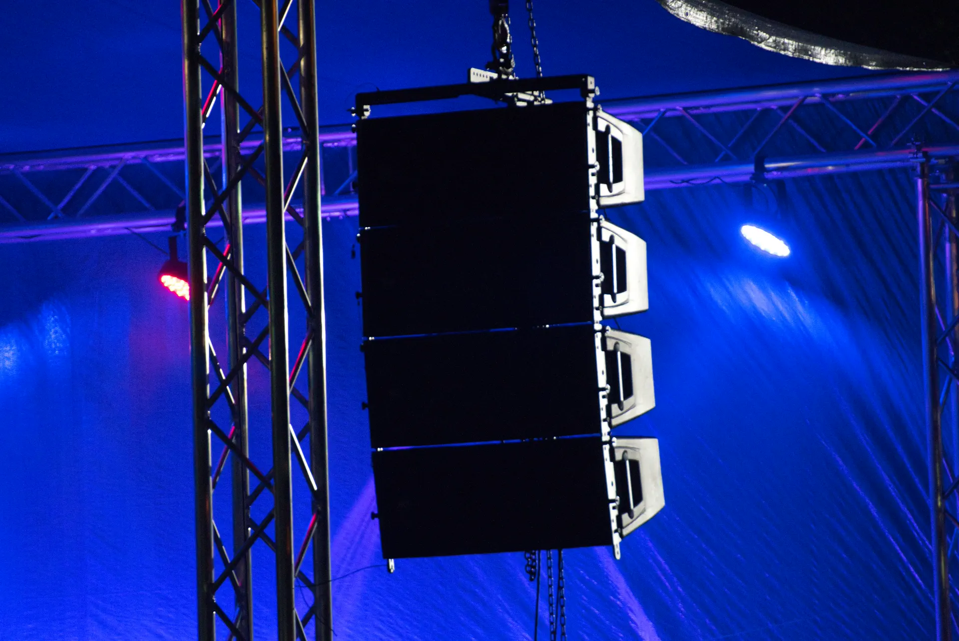 Racimo de cajas de line array colgado de un truss, recortado contra luces azules de escenario